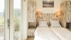 Une chambre avec deux lits, des têtes de lit en bois, des lampes de chevet, une vue à travers une porte vitrée et un tableau au mur.