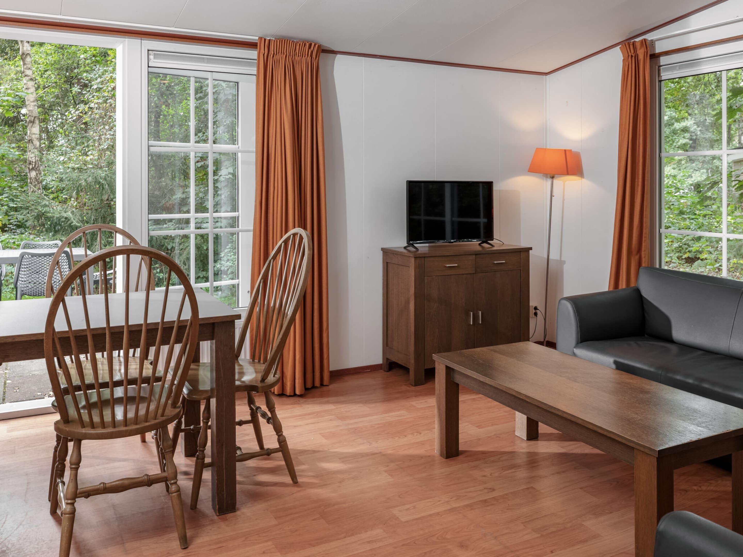 Una habitación con una mesa de comedor de madera, dos sillas de madera, un sofá, un televisor sobre un mueble bajo y una lámpara.