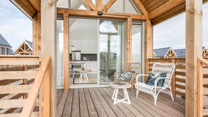 A wooden deck with two white wicker chairs and a small table, overlooking a modern kitchen and dining area through large glass doors.