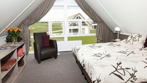 A bedroom with a bed, a chair, a small table with a vase of flowers, and a view of a house through a window. - Spacious Holiday Home With Garden (Tynaarlo)