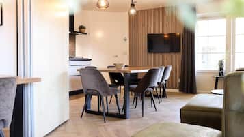 Una sala de estar moderna con una mesa de comedor de madera, sillas tapizadas en gris, un televisor de pantalla plana montado en una pared de madera y un ventanal con cortinas.