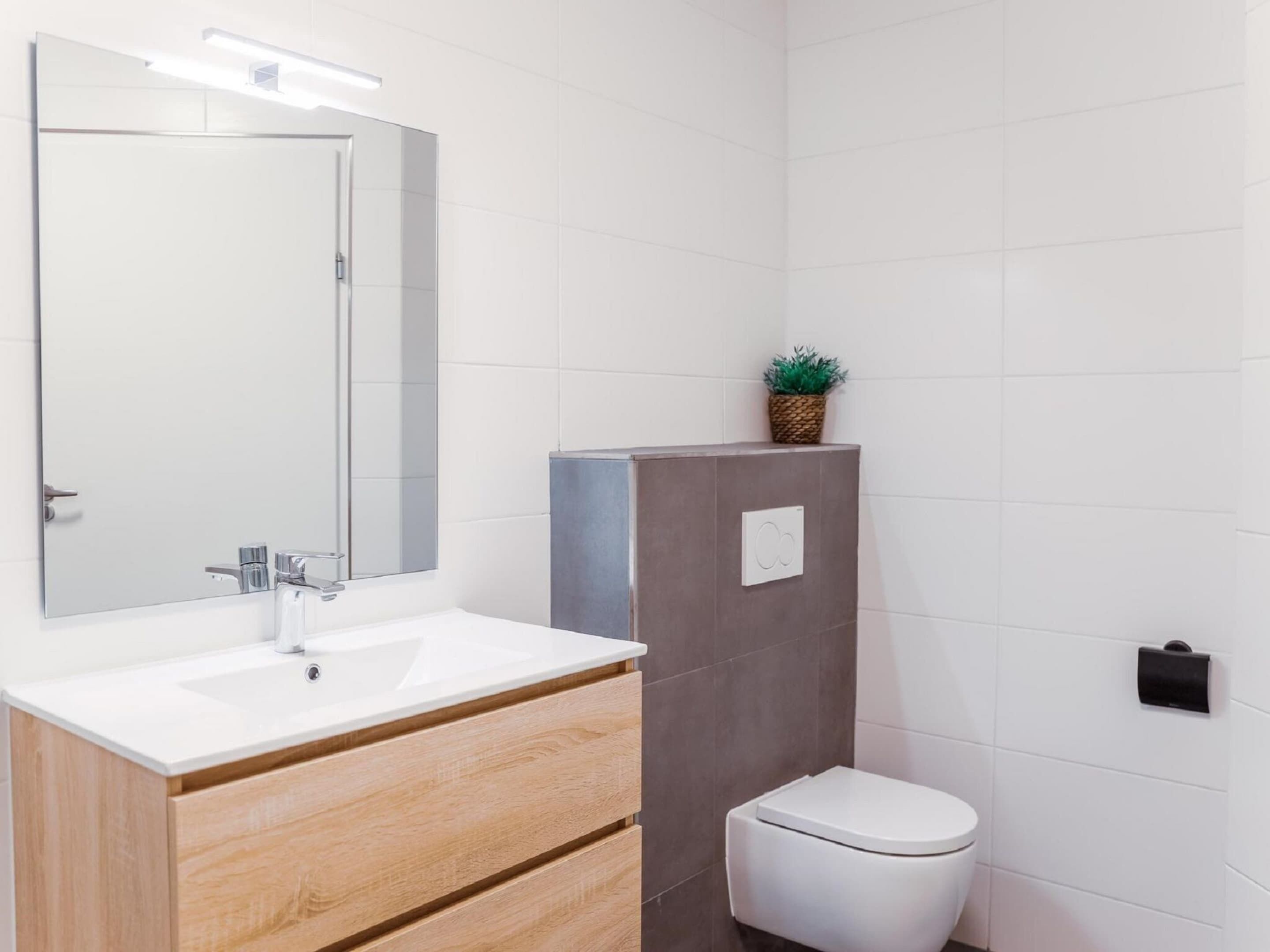Un baño moderno con un lavabo blanco, tocador de madera, espejo, inodoro y una pequeña planta en maceta.