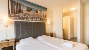 Habitación de hotel con una cama grande, cabecera, mesita de noche, lámpara, un cuadro en la pared y un baño visible a través de una puerta abierta.
