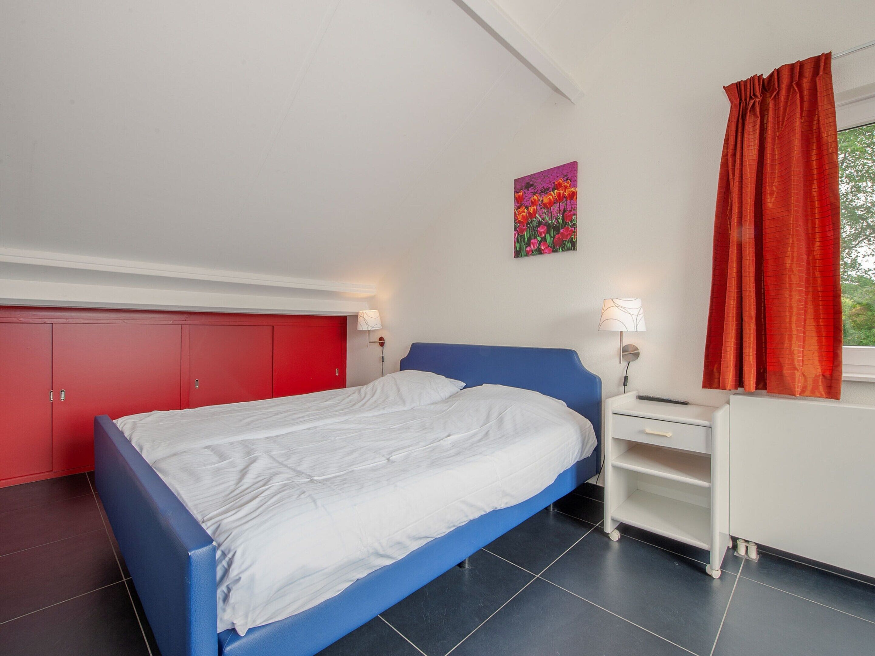 A bedroom with a blue bed, white walls, a red wardrobe, and a painting of flowers.