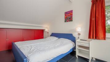A bedroom with a blue bed, white walls, a red wardrobe, and a painting of flowers.