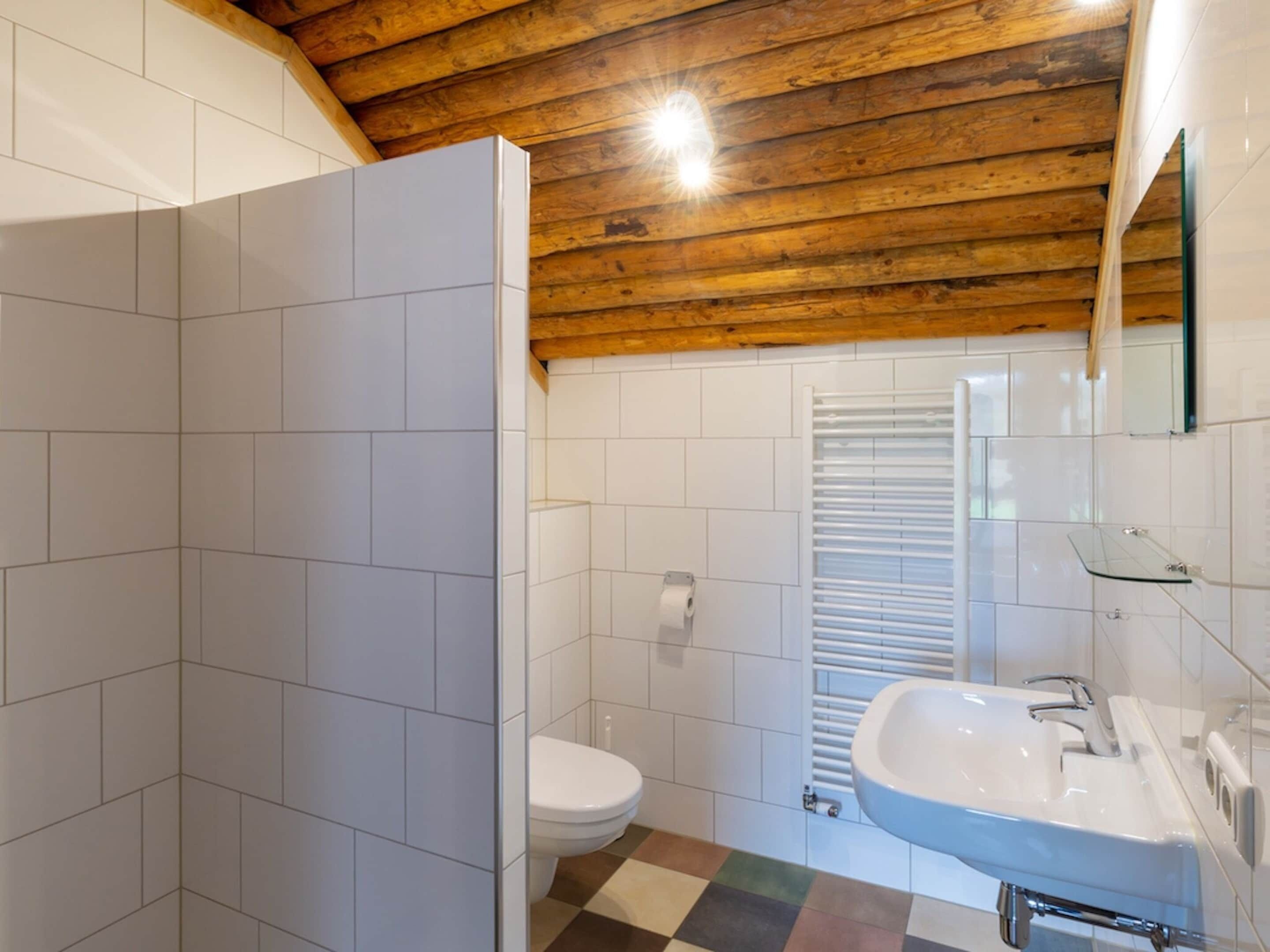 Une salle de bain avec des carreaux blancs, un plafond en bois, des toilettes, une douche en verre et un lavabo avec un miroir.