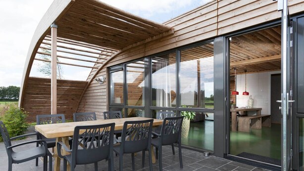 Un espace de restauration en plein air avec une table et des chaises en bois, de grandes portes-fenêtres coulissantes donnant sur un espace intérieur, et une structure en bois incurvée au-dessus.