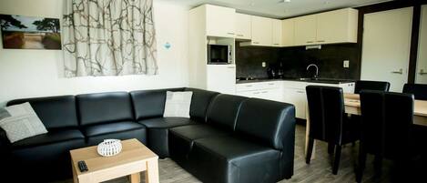 Una sala moderna con un sofá de cuero negro, una mesa de centro de madera y una zona de comedor con una mesa y sillas negras. Se ve una cocina con armarios blancos y encimera negra al fondo.