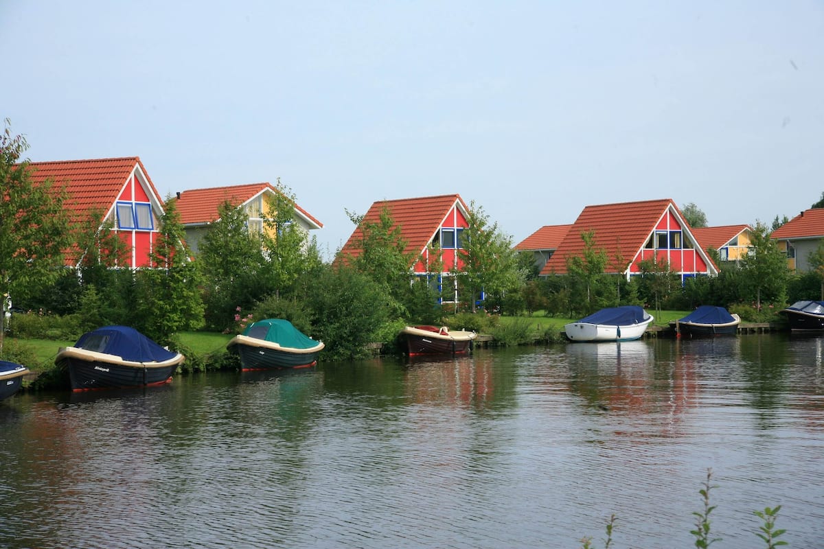 Eine Reihe von Häusern mit roten Dächern an einem ruhigen Gewässer, vor dem Boote vor Anker liegen.