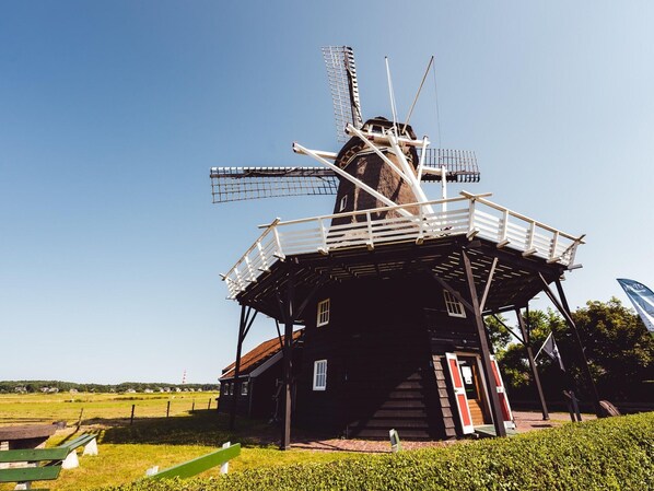 A windmill with a wooden structure and a metal roof, surrounded by a grassy area and a paved path. - Wooden Bungalow With Dishwasher Near the Beach (Hollum)
