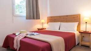 Ein ordentlich bezogenes Bett mit einer roten und beigen Tagesdecke, zwei Nachttischlampen und einem Fenster mit Vorhängen.