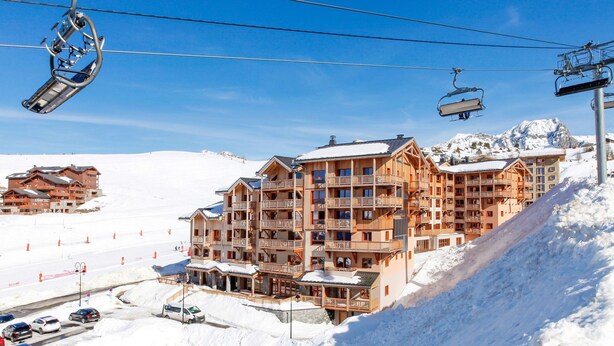 Um hotel com vários andares, varandas, teleféricos e uma montanha nevada ao fundo.