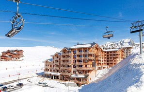 Un hotel de varios pisos con balcones, telesillas y una montaña nevada al fondo.
