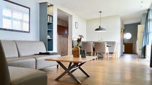 A modern living room with a sofa, a wooden dining table, and a kitchen in the background. - Bungalow in Netherlands With Dune Garden ('s-Gravenzande)
