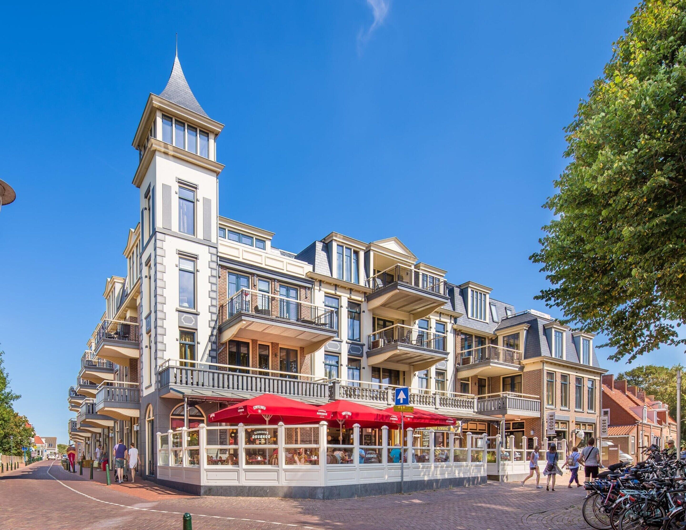 Een meerlaags gebouw met een opvallende toren, balkons en buitenzitplaatsen met rode parasols.