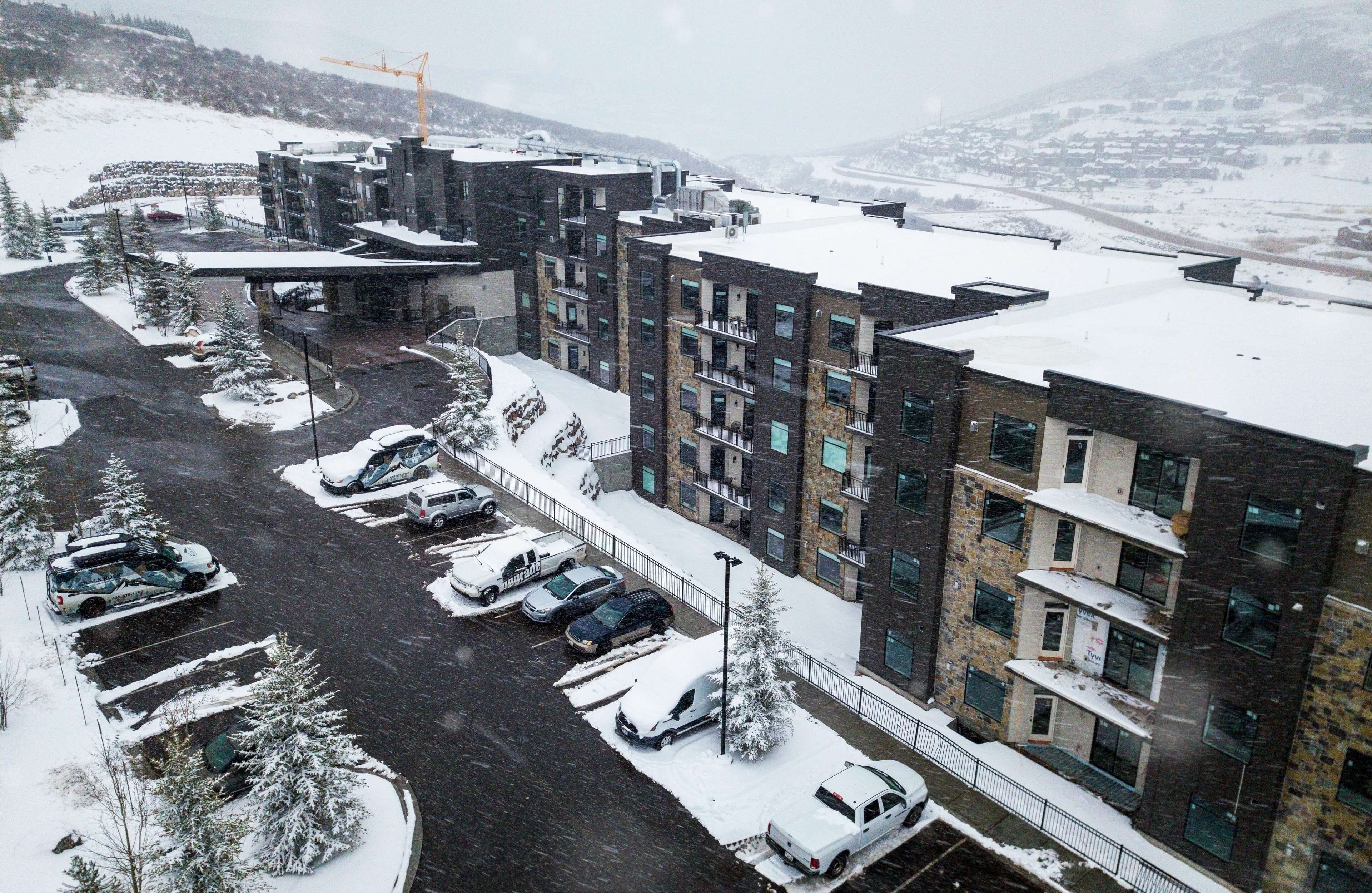 A multi-story building with a parking lot in front, surrounded by snow-covered trees and a mountainous backdrop.