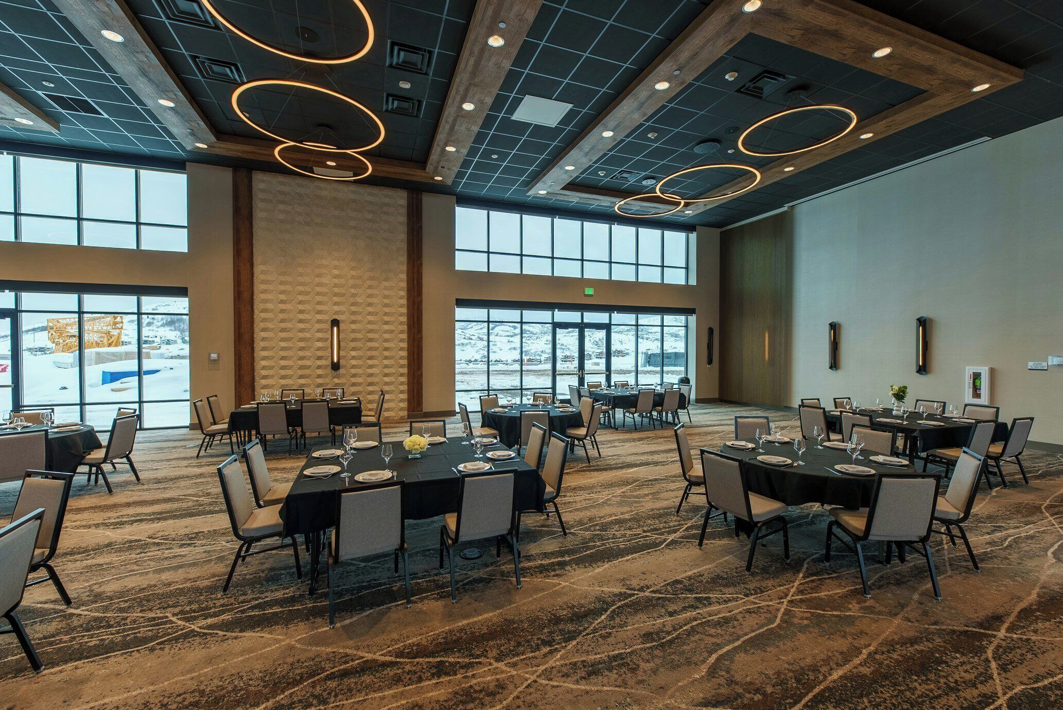 A spacious conference room with multiple tables set for dining, large windows offering a view of a snowy landscape, and a modern ceiling design with circular light fixtures.