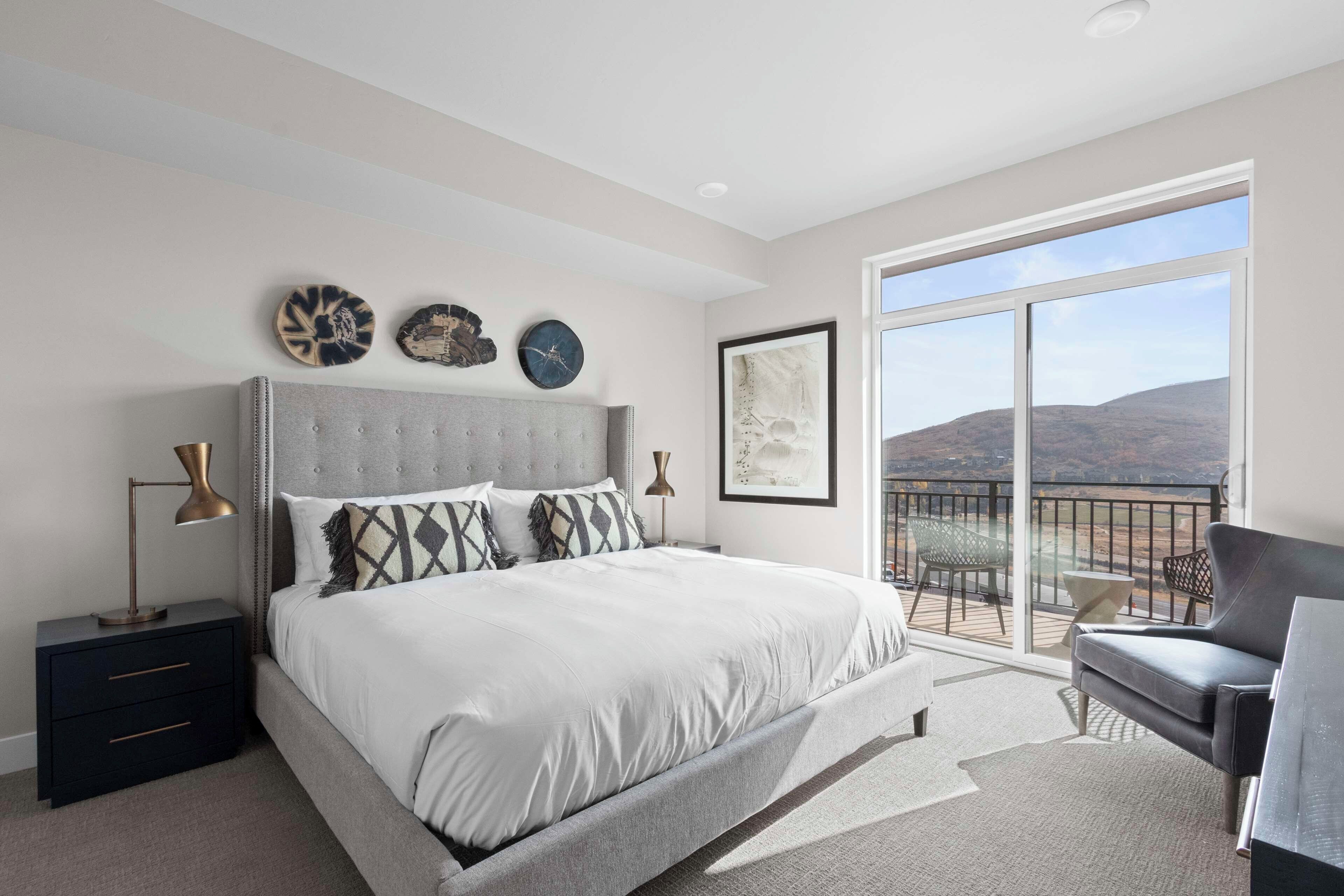 A bedroom with a large bed, a nightstand, a desk, and a chair. There is a balcony with a view of the hills.