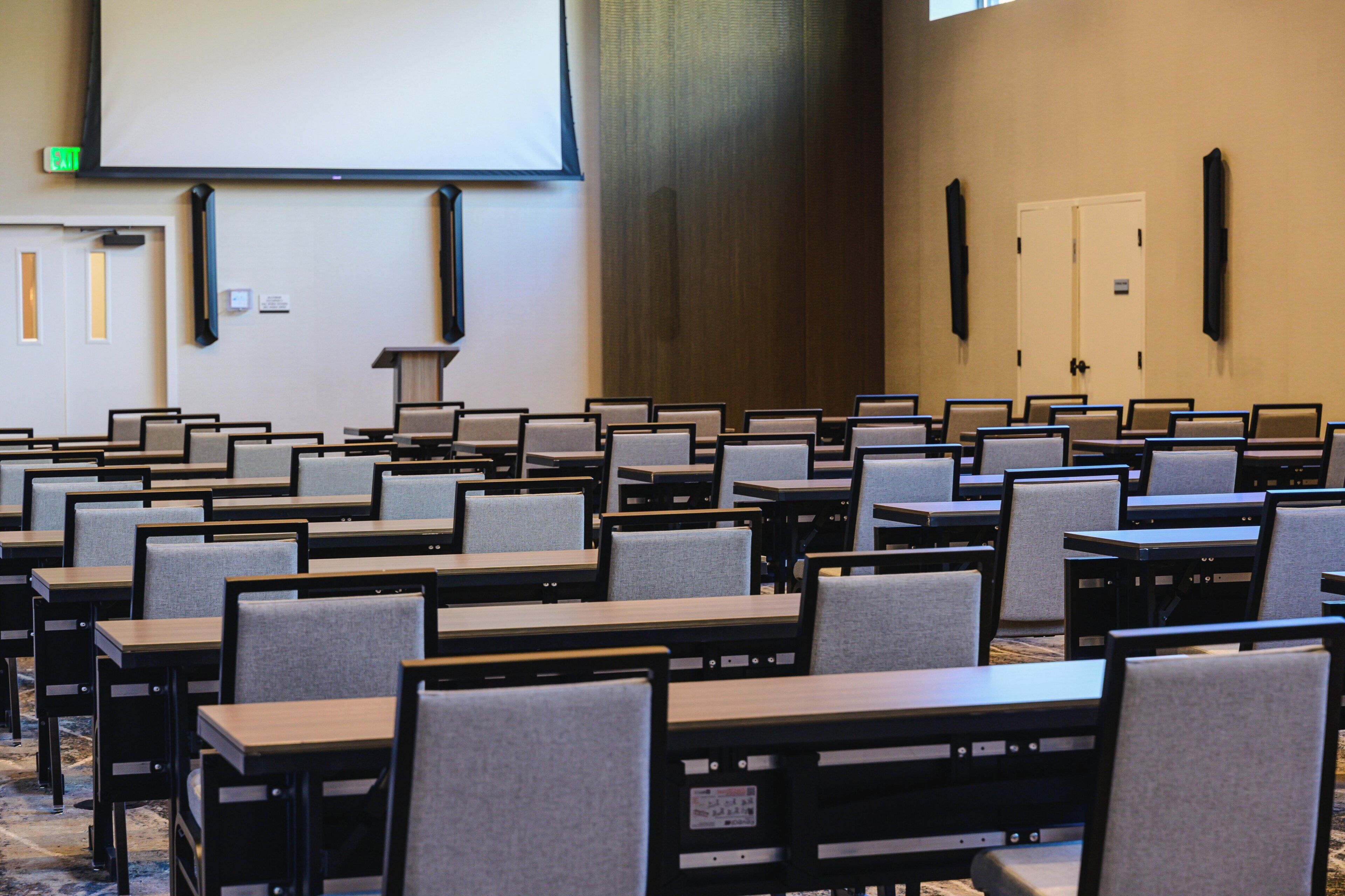 A conference room with rows of tables and chairs, a projector screen, and a door with a sign.