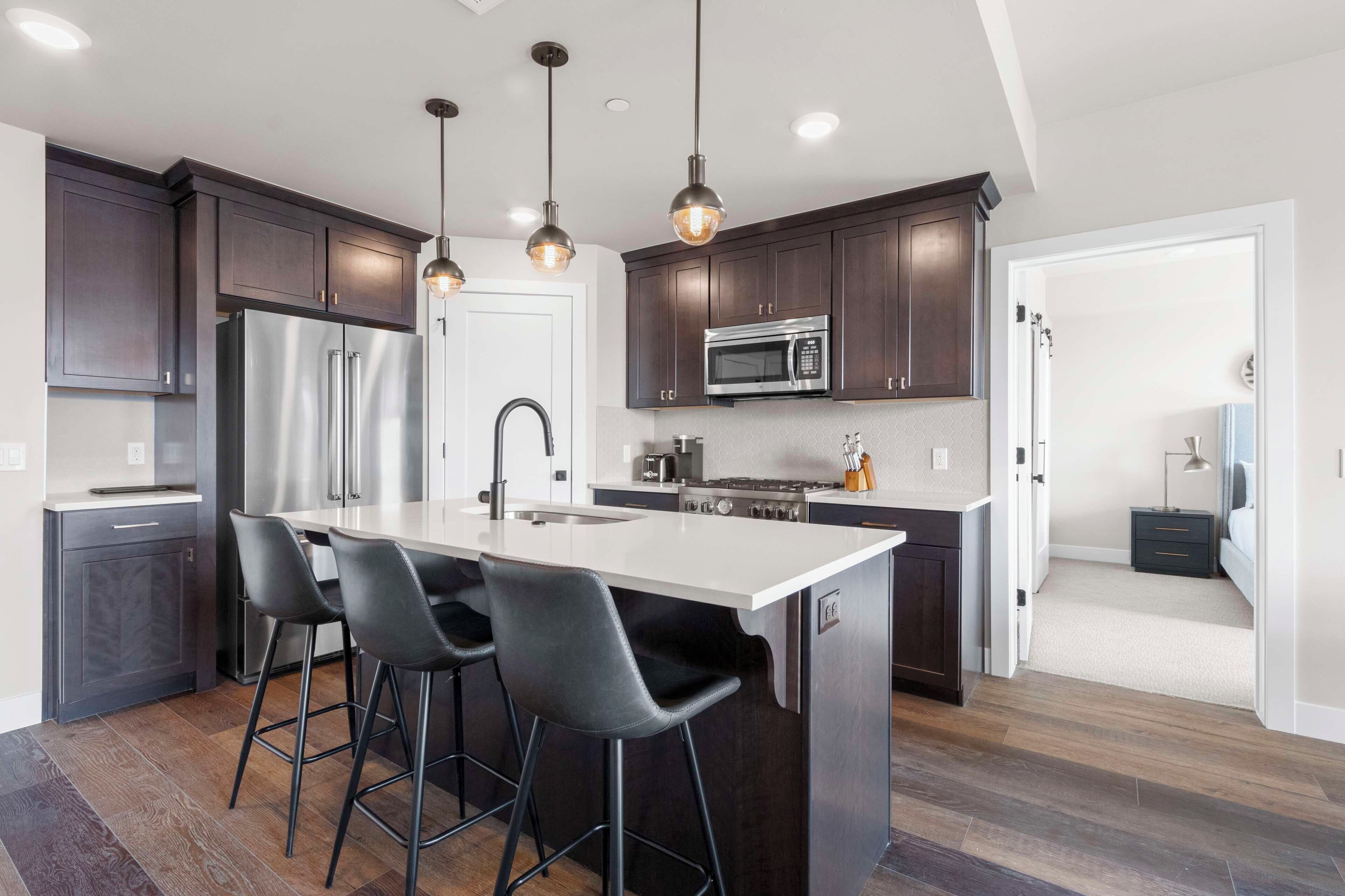 A modern kitchen with dark wood cabinets, stainless steel appliances, a central island with bar stools, and pendant lighting.