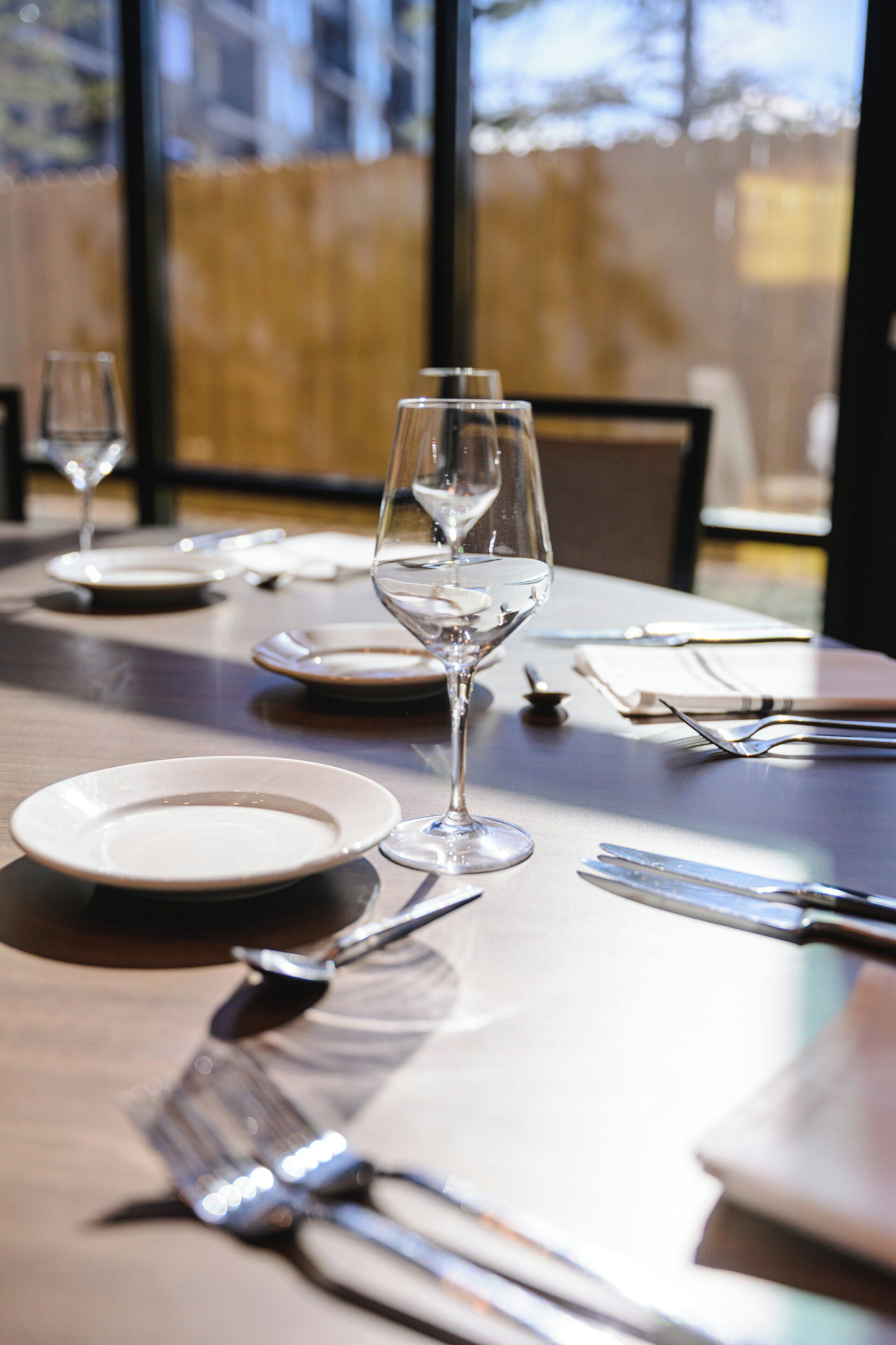 A dining table set with plates, cutlery, and wine glasses, with a view of the outdoors through a window.