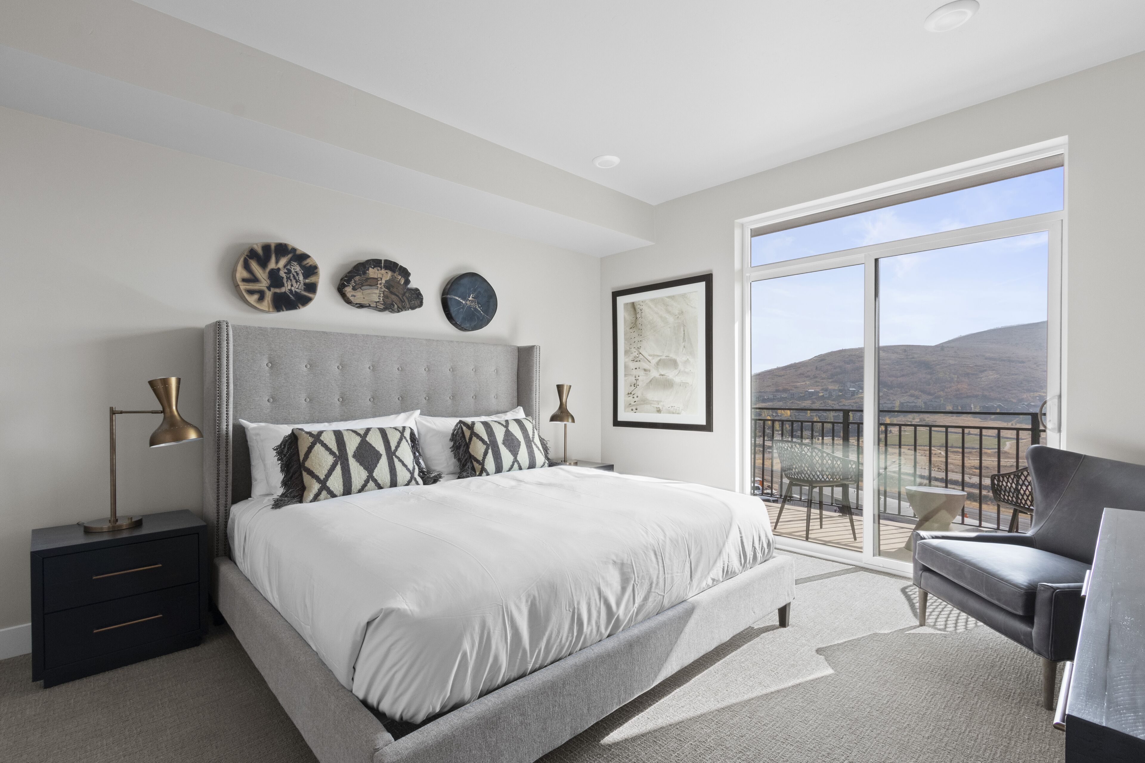 A bedroom with a large bed, a nightstand, a desk, and a balcony with a view of the hills.