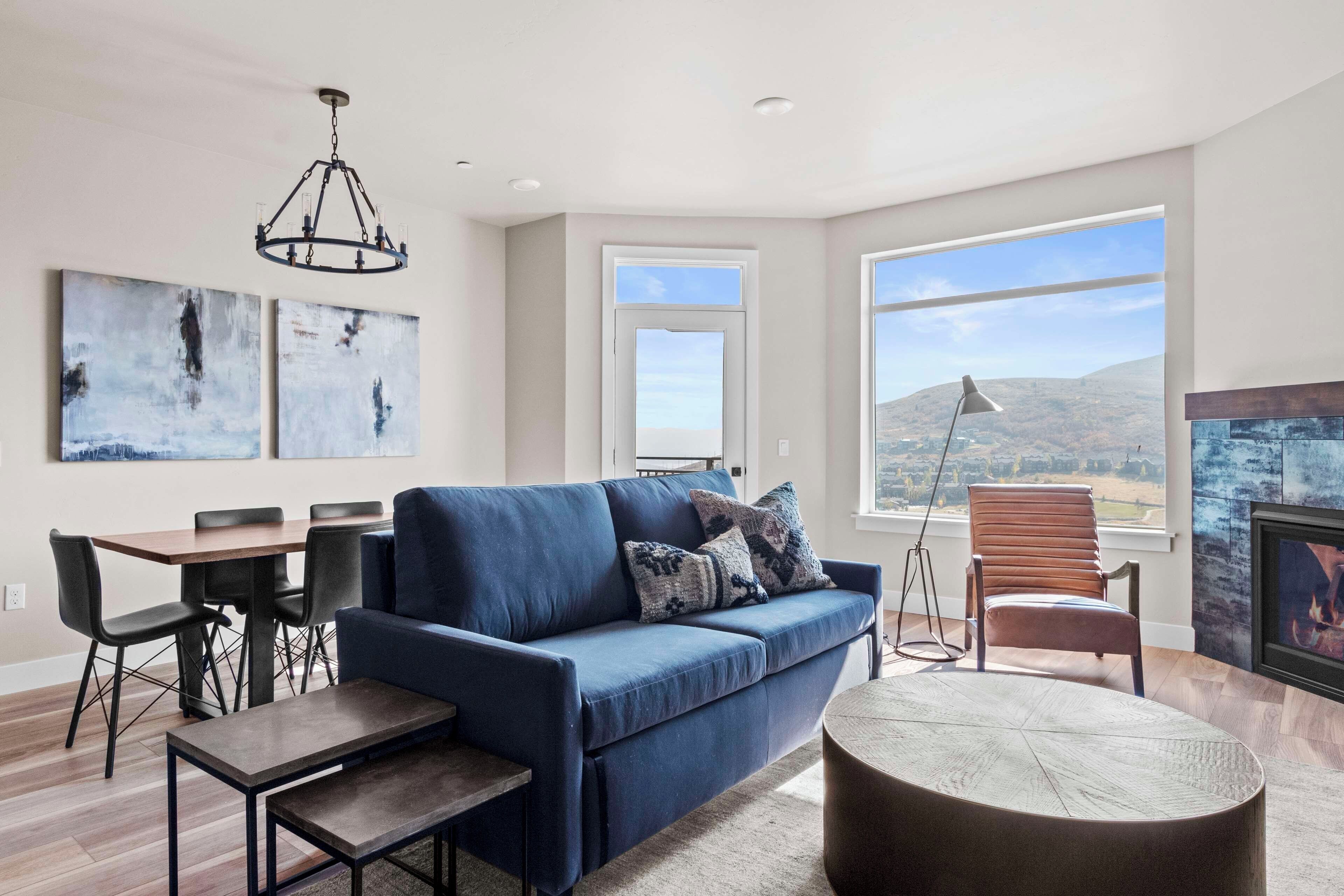 Una sala moderna con un sofá azul, una mesa de comedor con sillas, una mesa redonda de centro, una chimenea y ventanas amplias con vistas al paisaje.