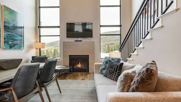 A modern living room with a fireplace, a flat-screen TV mounted above it, a dining area with a table and chairs, and a staircase with a railing.
