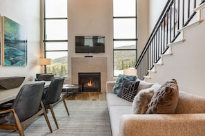 A modern living room with a fireplace, a flat-screen TV mounted above it, a dining area with a table and chairs, and a staircase with a railing.