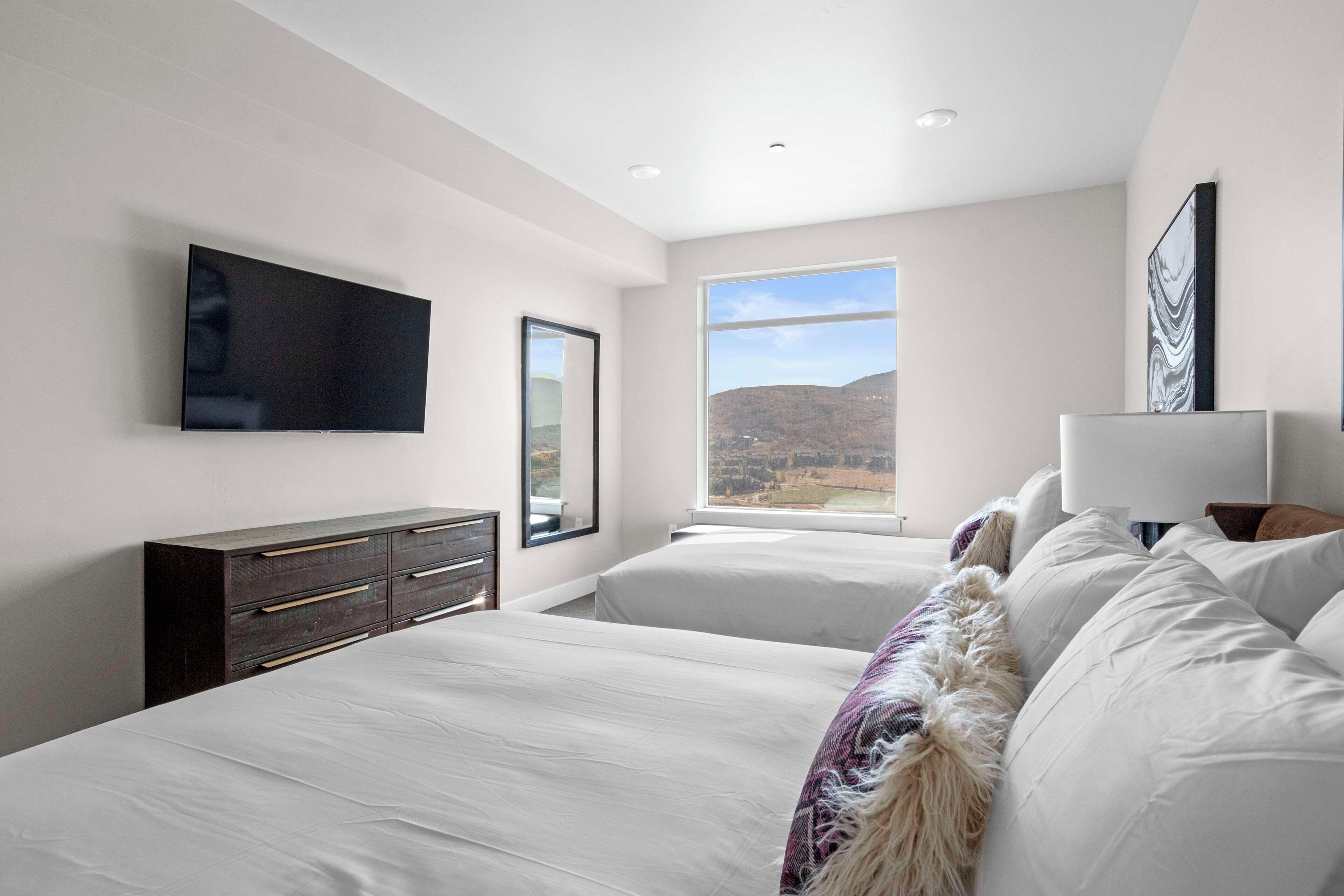 A bedroom with two beds, a TV, a dresser, a window with a view, and a painting.