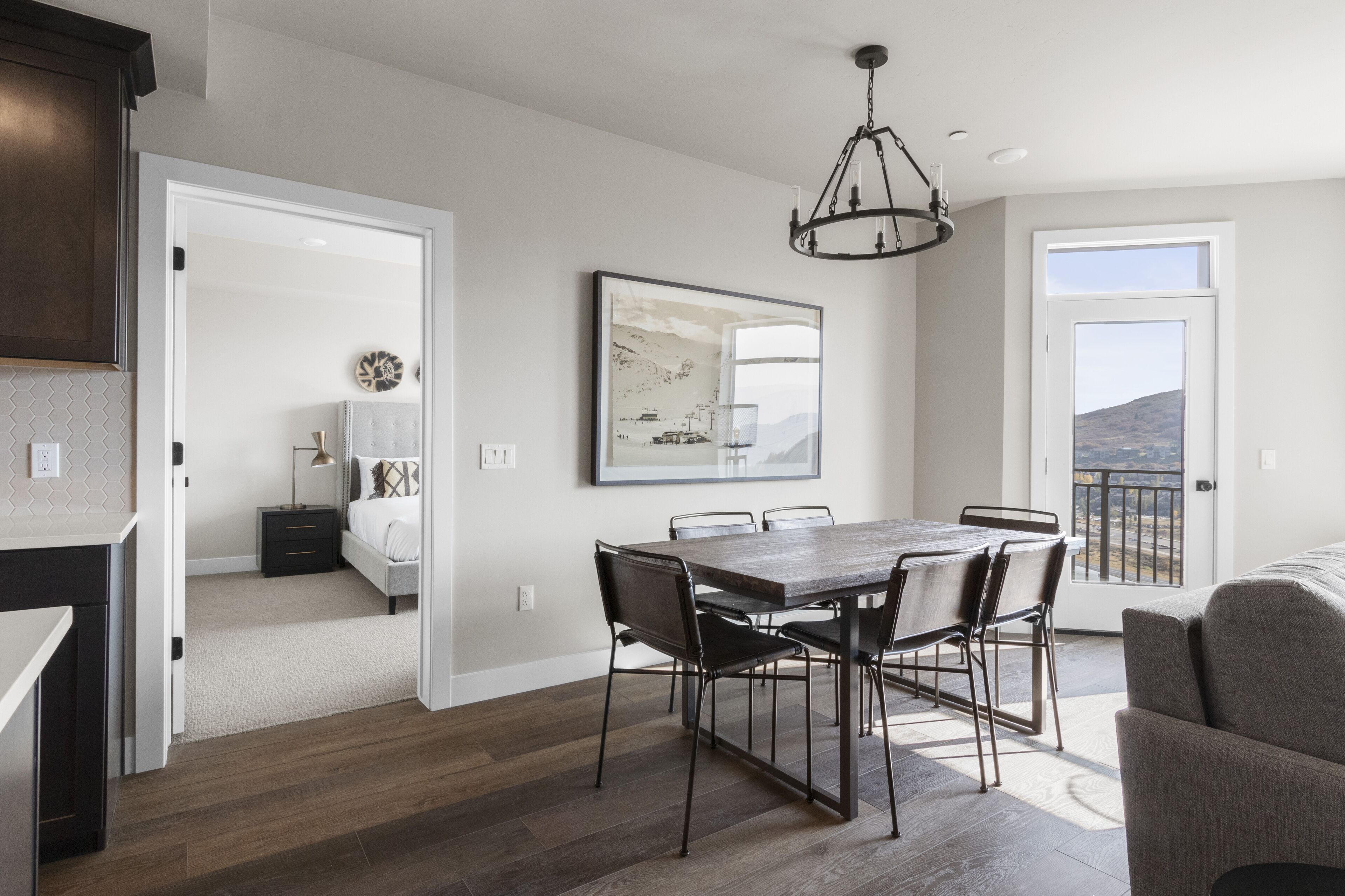 Una cocina moderna con piso de madera oscura, un comedor con una mesa grande y sillas, una lámpara de candelabro y vistas a través de una puerta.