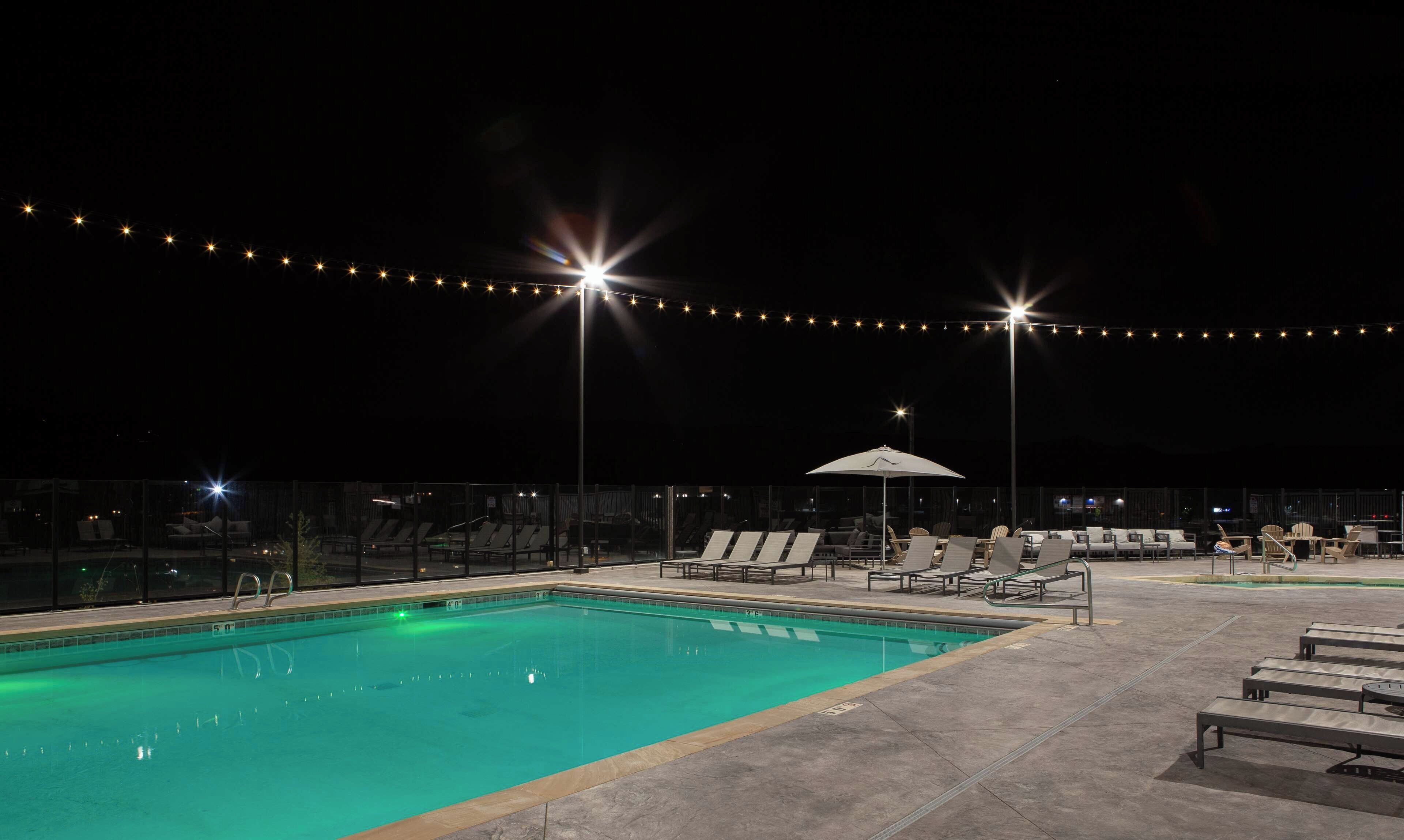 A swimming pool with lounge chairs and umbrellas, surrounded by a fence and string lights.