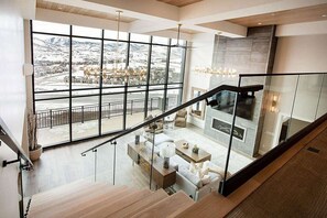 Un interior moderno con una escalera que lleva a un espacio de estar con chimenea, y grandes ventanales que ofrecen vistas de un paisaje nevado.