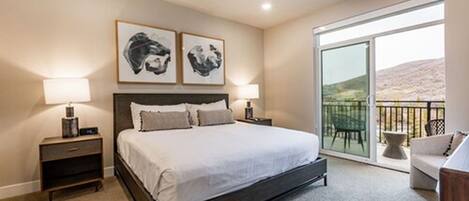 A bedroom with a large bed, two bedside tables, a nightstand, a balcony with a view, and two framed artworks on the wall.