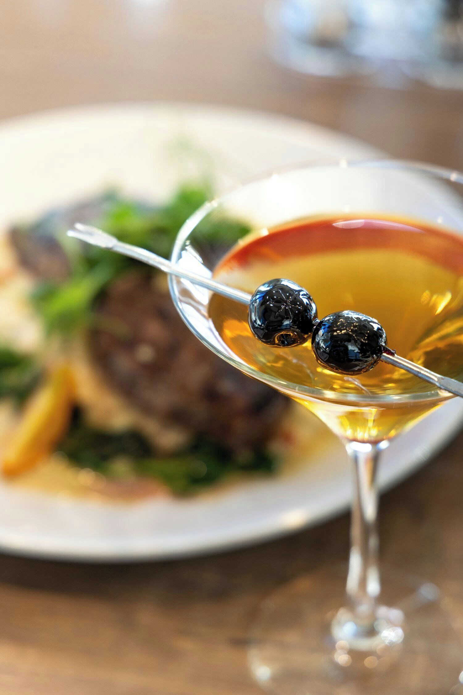 Una copa de martini con pajita y dos aceitunas en un palillo, frente a un plato con comida.