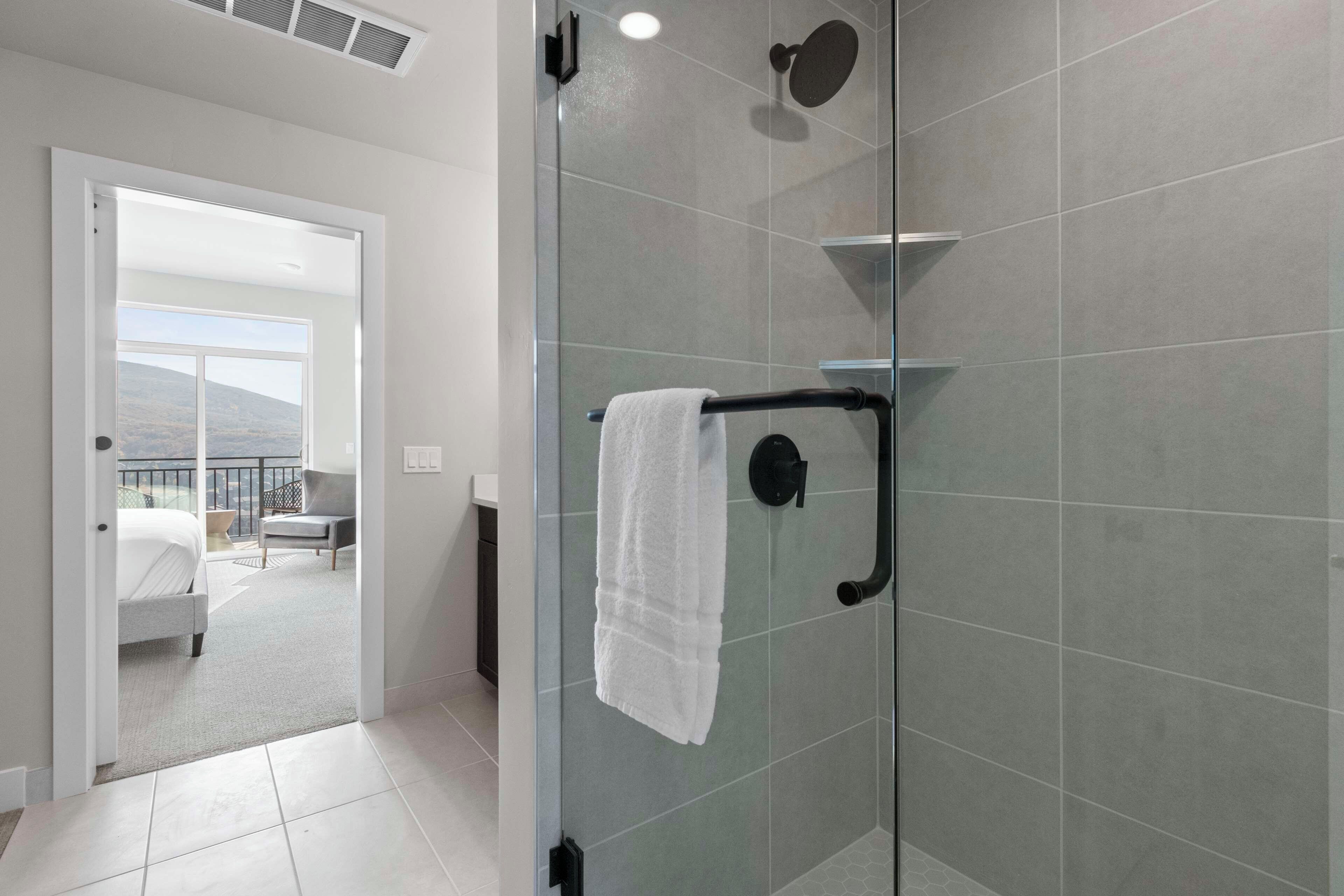 Ein modernes Badezimmer mit Duschkabine aus Glas, einem Handtuchhalter und Blick auf einen Balkon mit Bett und Stuhl.