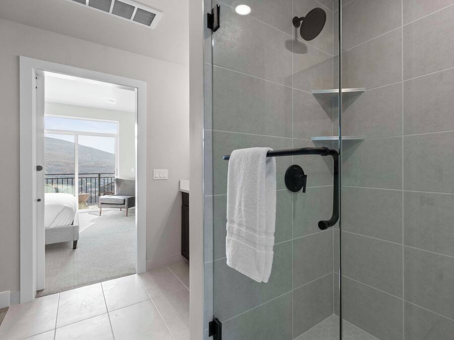 A modern bathroom with a glass shower enclosure, a towel rack, and a view of a balcony with a bed and a chair.