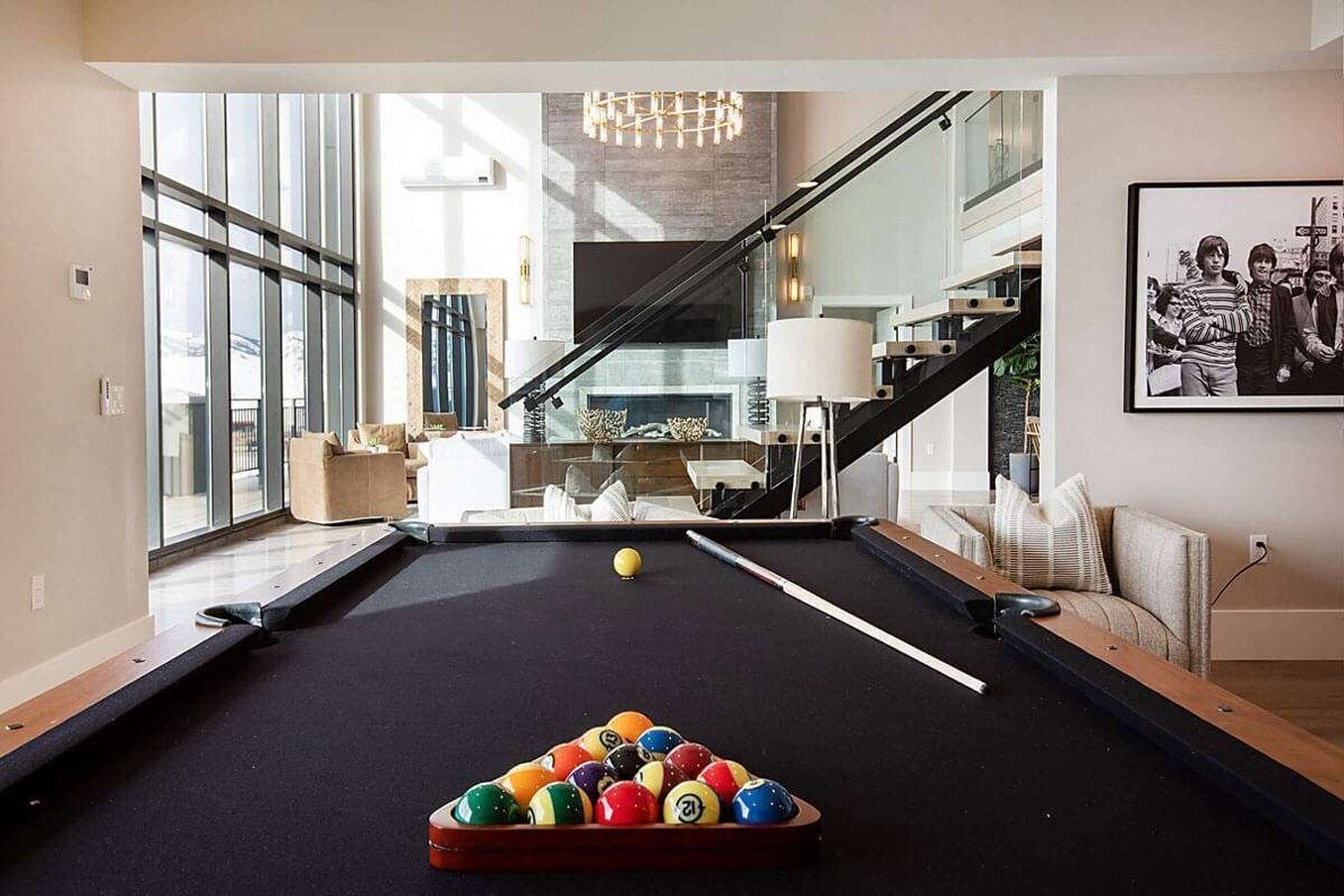 A modern living room with a pool table, a staircase, and a large window.