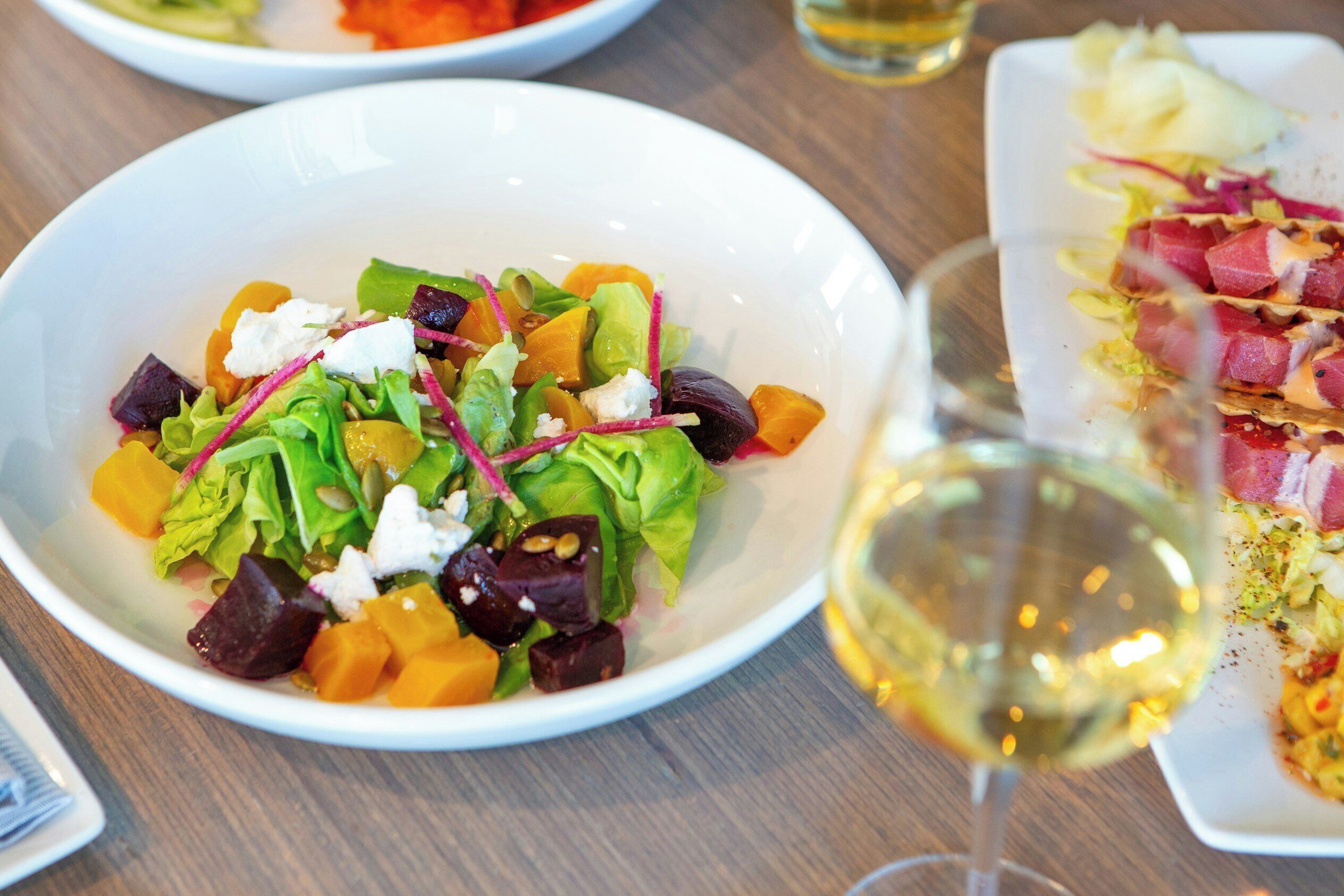 A table with a bowl of salad, a glass of white wine, and a plate with food.