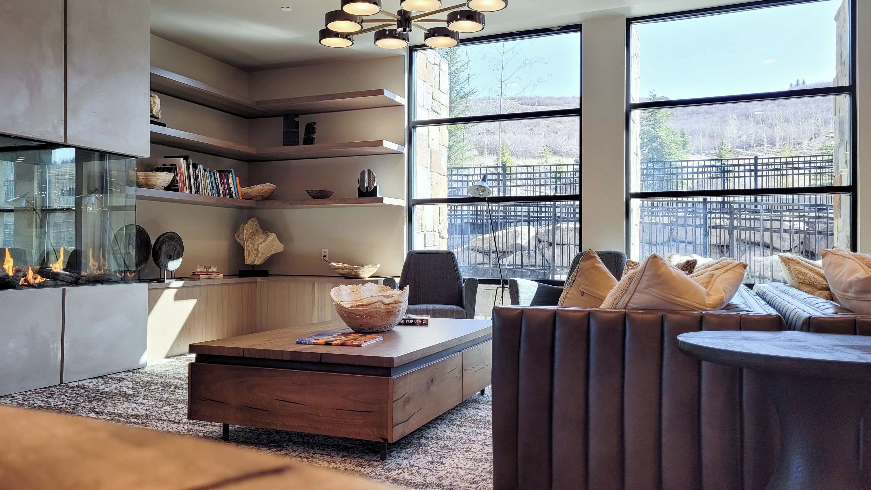 A modern living room with a fireplace, bookshelves, a sofa, and large windows offering a view of the outdoors.