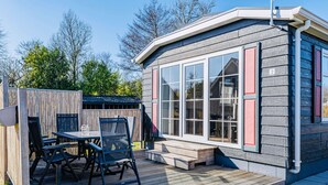 Een klein, modern huis met een terras, een tafel met stoelen en een groot raam.