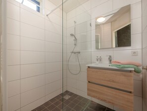 Un baño moderno con cabina de ducha de vidrio, azulejos blancos, tocador de madera y espejo con luz superior.