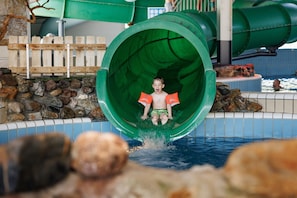 Un bambino scivola lungo uno scivolo acquatico verde in una zona con piscina, rocce e una recinzione in legno sullo sfondo.