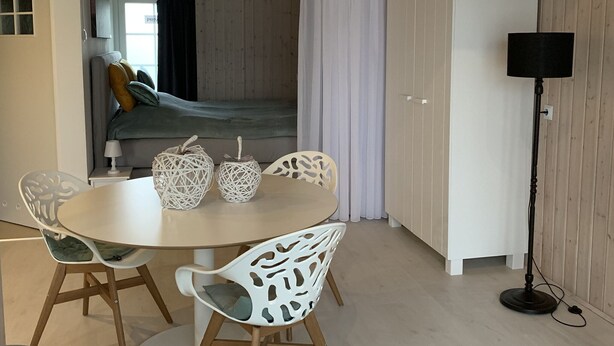 Une pièce avec une table ronde, des chaises blanches, un lit avec une tête de lit grise, une armoire et un lampadaire.
