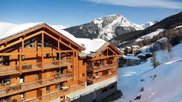 Ein mehrstöckiges Holzchalet mit Balkonen vor einer verschneiten Berglandschaft.