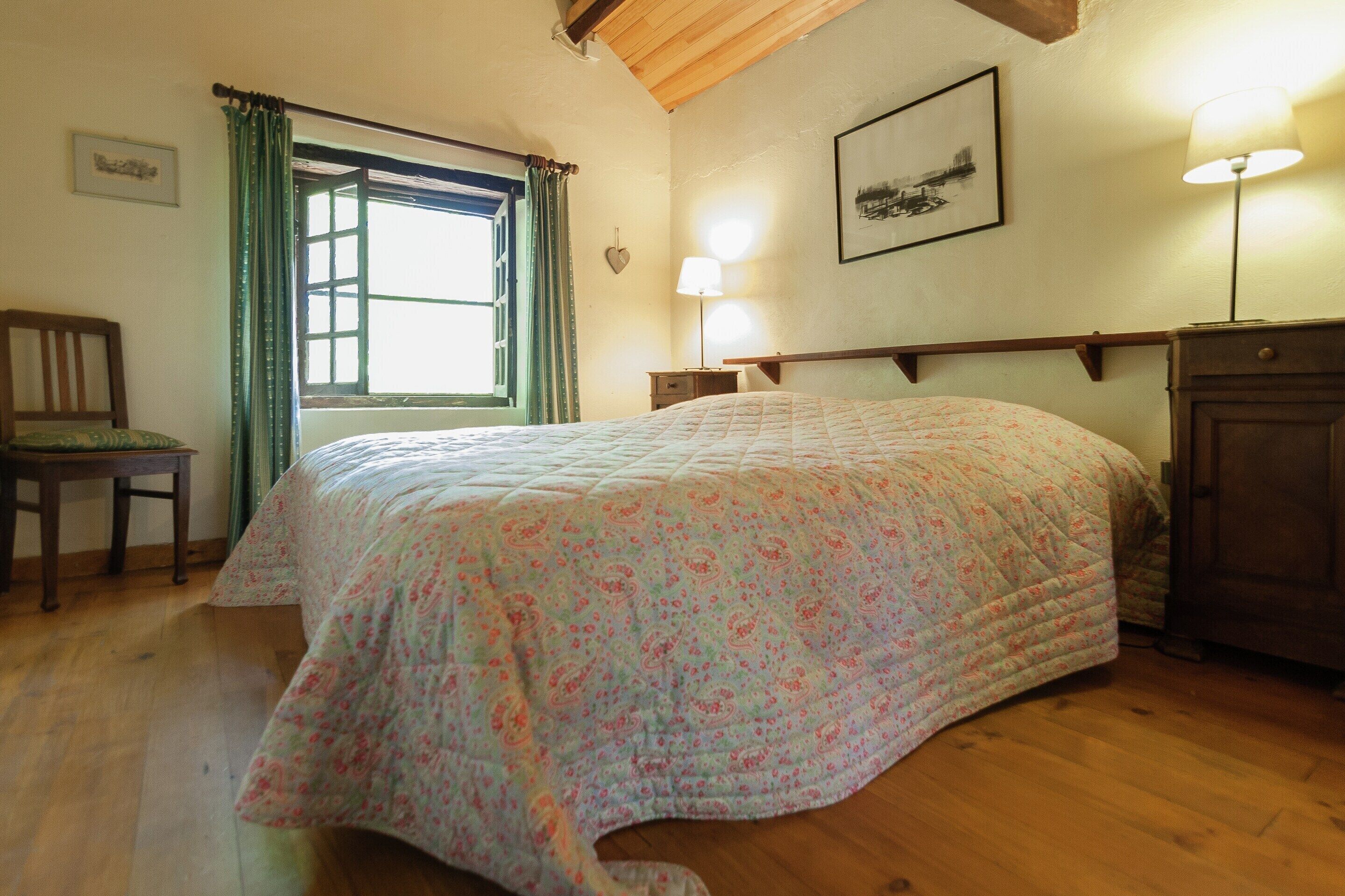 Un dormitorio con cama, una silla, ventana con cortinas, mesita de noche, lámpara y un cuadro en la pared.