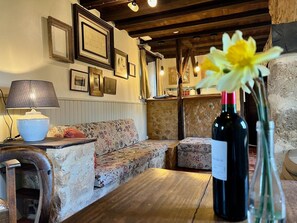 A cozy room with a wooden table, a sofa, and a lamp. There are framed pictures on the wall and a bottle of wine on the table. - Cottage in Vezere Valley Near Lascaux Caves (Les Eyzies)