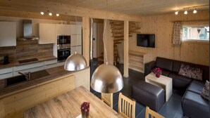 Moderni keittiö, jossa on puiset kaapit, ruokapöytä tuoleineen, sohva ja seinälle asennettu televisio.