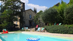 Une piscine avec un tapis flottant, entourée de chaises longues et d’un bâtiment en pierre.