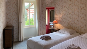 A bedroom with floral wallpaper, a window with curtains, a bed with pillows, a bedside table with a lamp, and a wooden cabinet.